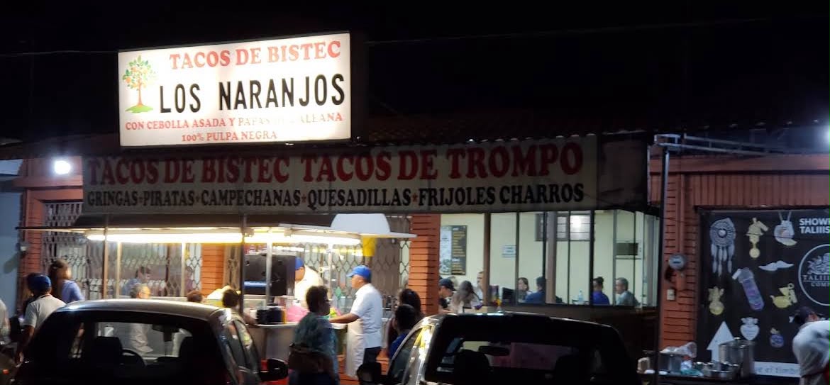 Letrero de Tacos de Bistec Los Naranjos con clientes en la banqueta